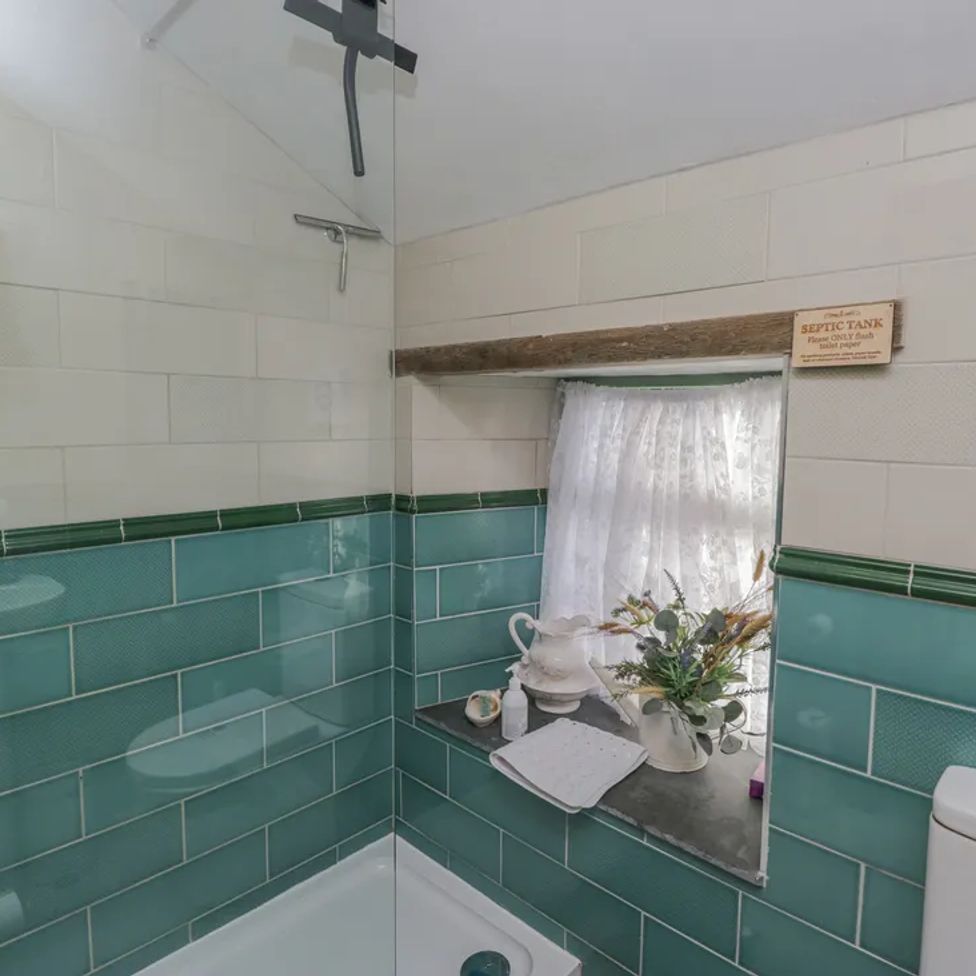A bathroom with a shower area green and white tiled walls a small window with a curtain flowers and toiletries on the window sill and a toilet at The Old Cart House in Garsdale near Sedbergh