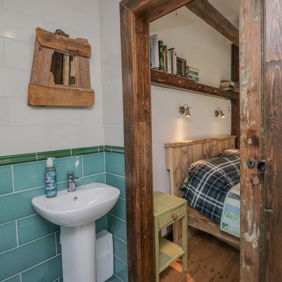 A bathroom with a sink and mirror next to a wooden door opening to a bedroom with a bed and bookshelf at The Old Cart House in Garsdale near Sedbergh