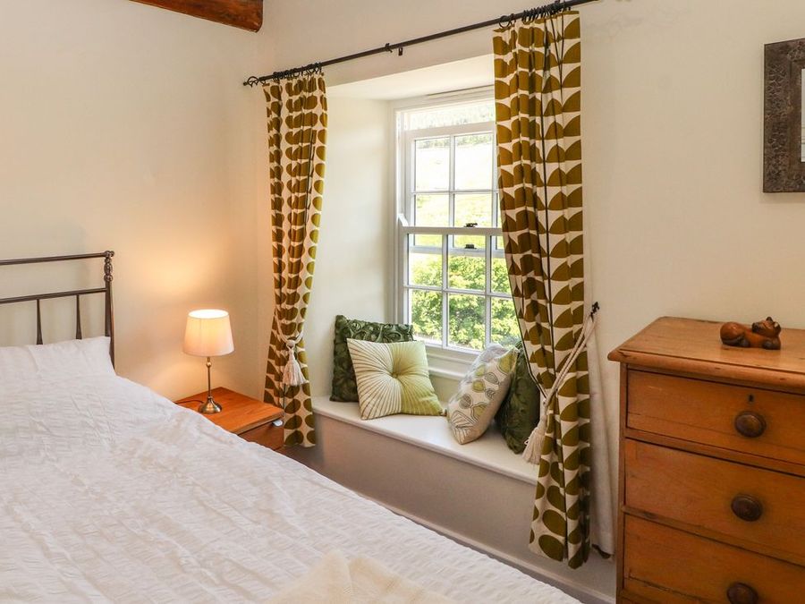 A bedroom with a bed a bedside table with lamp a window seat with cushions patterned curtains and a wooden chest of drawers at Roger Pot in Garsdale near Sedbergh