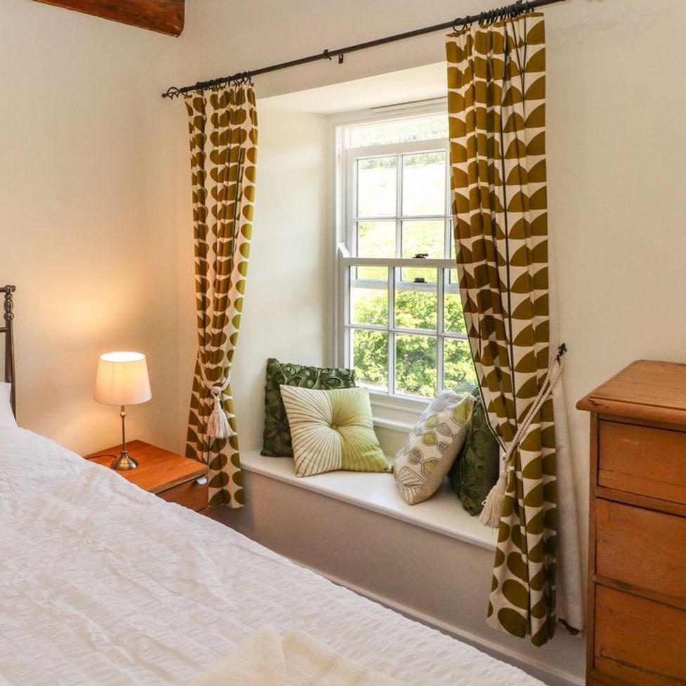 A bedroom with a bed a bedside table with lamp a window seat with cushions patterned curtains and a wooden chest of drawers at Roger Pot in Garsdale near Sedbergh