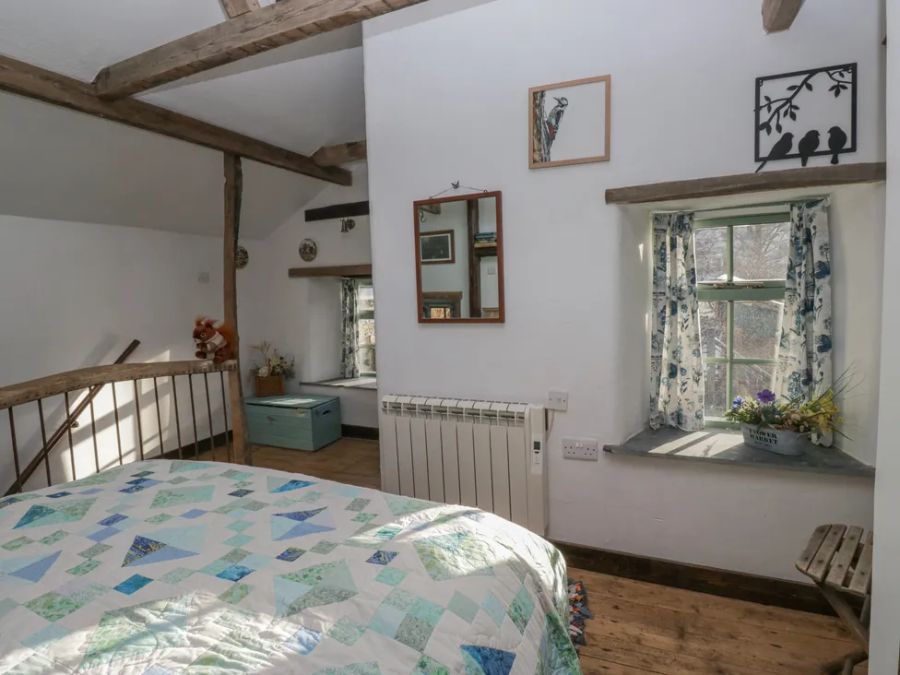 A bedroom with a bed a window with floral curtains a mirror on the wall and wooden beams at The Old Cart House in Garsdale near Sedbergh