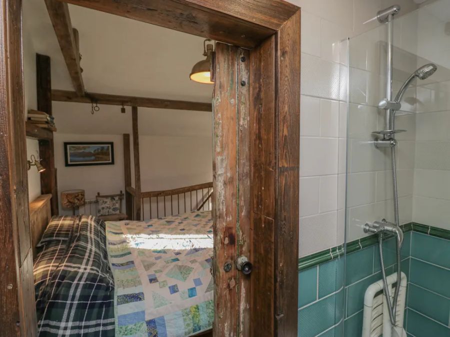 A bedroom with a bed and chair visible through a wooden door next to a shower area with green and white tiles at The Old Cart House in Garsdale near Sedbergh