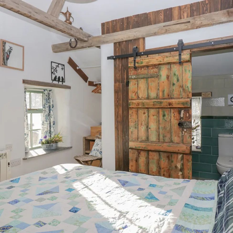 A bedroom with a patterned quilt on the bed a wooden sliding door leading to a bathroom and a window with floral curtains at The Old Cart House in Garsdale near Sedbergh