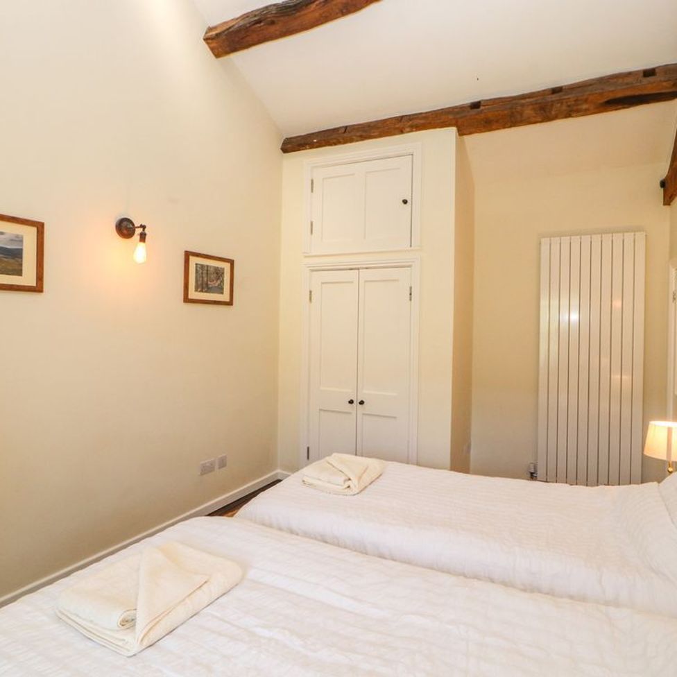 A bedroom with two single beds with white bedding towels on beds a wall light framed pictures a lamp a wardrobe and exposed wooden beams at Roger Pot in Garsdale near Sedbergh