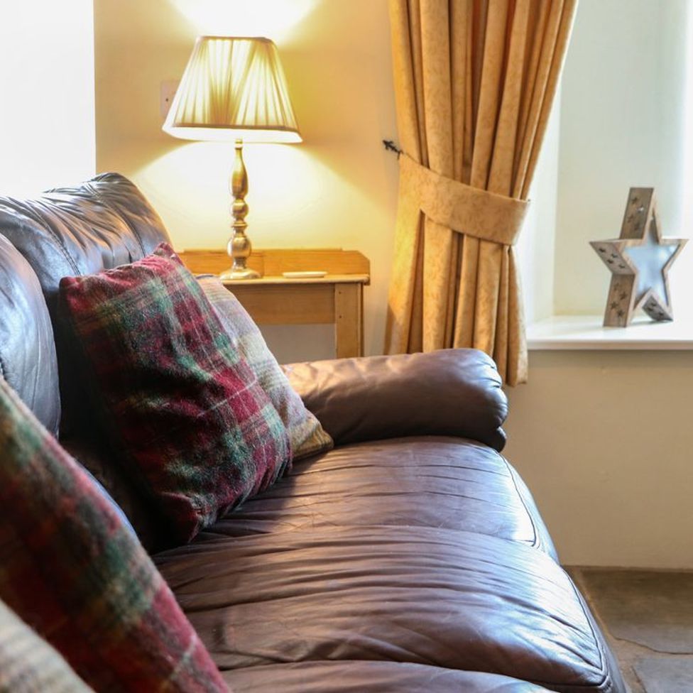 A leather sofa with plaid cushions next to a wooden side table with a lamp and yellow curtains with star decorations on the windowsill at Roger Pot in Garsdale near Sedbergh