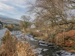 A river flowing alongside a road with leafless trees and a house in the background at The Old Cart House in Garsdale near Sedbergh