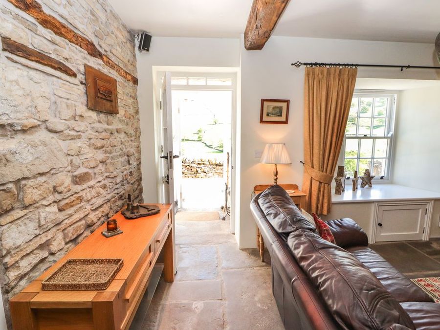 A room with a leather sofa near a window with curtains and a wooden console table against a stone wall at Roger Pot in Garsdale near Sedbergh