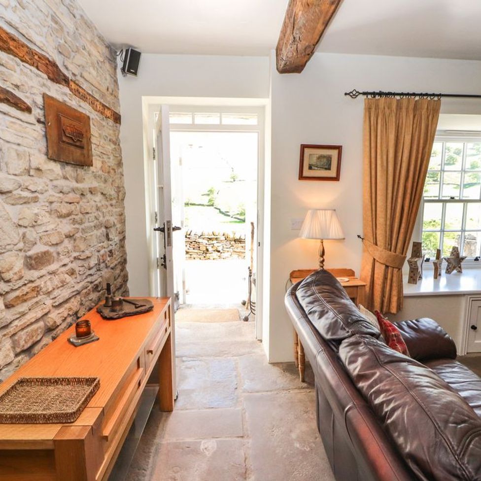 A room with a leather sofa near a window with curtains and a wooden console table against a stone wall at Roger Pot in Garsdale near Sedbergh