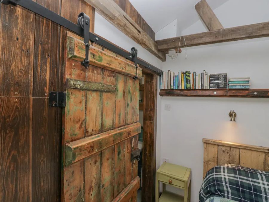 A rustic sliding wooden door next to a small bedside table and a shelf with books in a bedroom at The Old Cart House in Garsdale near Sedbergh