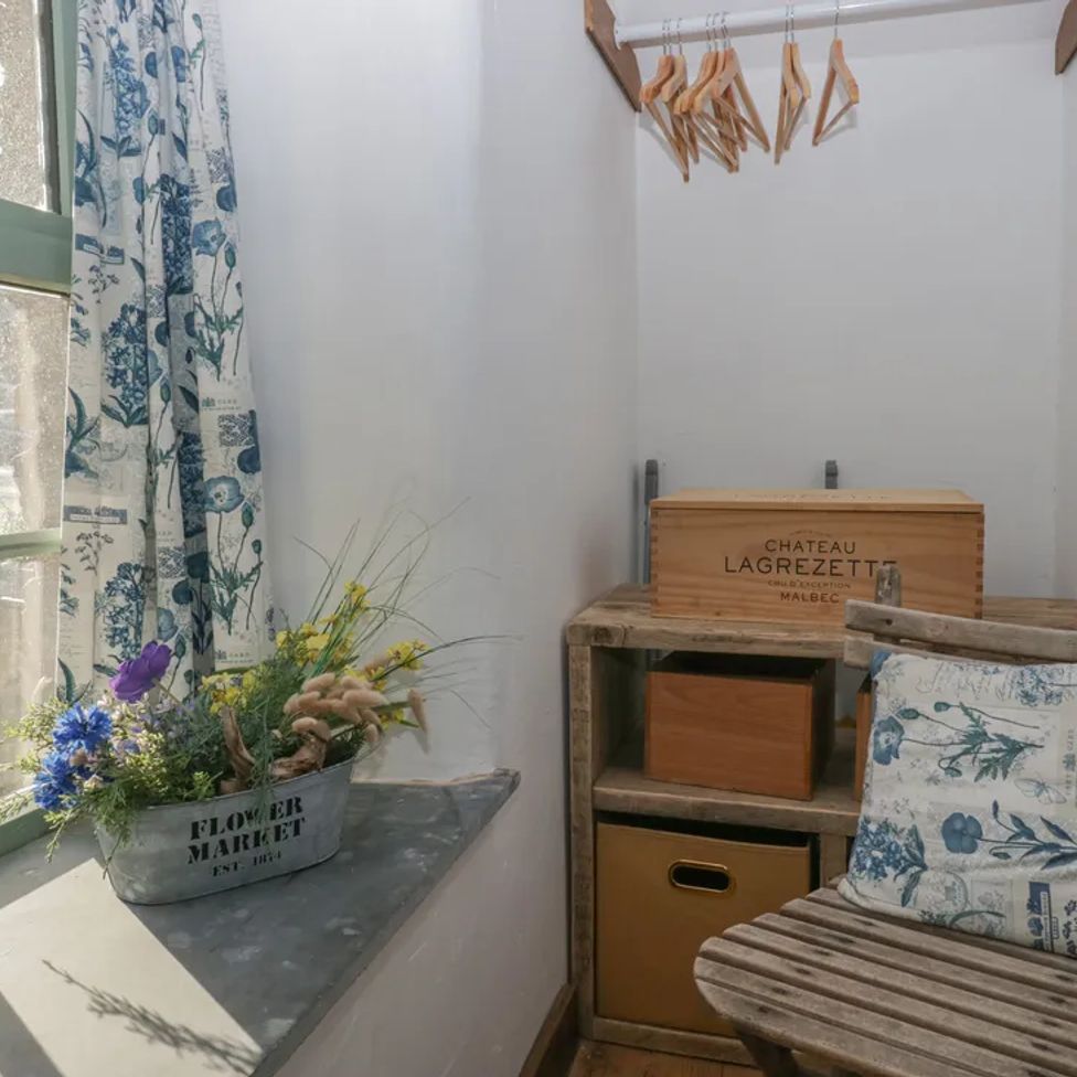 A small corner with a window, a flowerpot on the sill, a wooden chair with a cushion, hangers, and storage boxes at The Old Cart House in Garsdale near Sedbergh