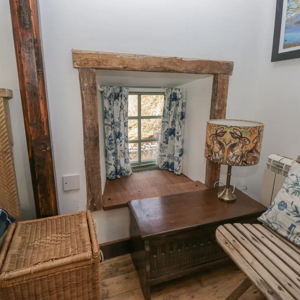 A small seating area with a wooden chair a wicker basket a wooden table and a window with curtains at The Old Cart House in Garsdale near Sedbergh