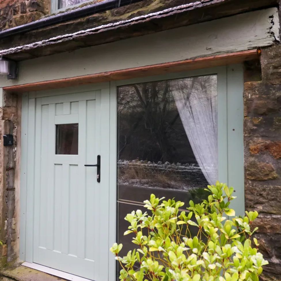 A stone building entrance with a light green door and window with curtains and plants near the door at The Old Cart House in Garsdale near Sedbergh