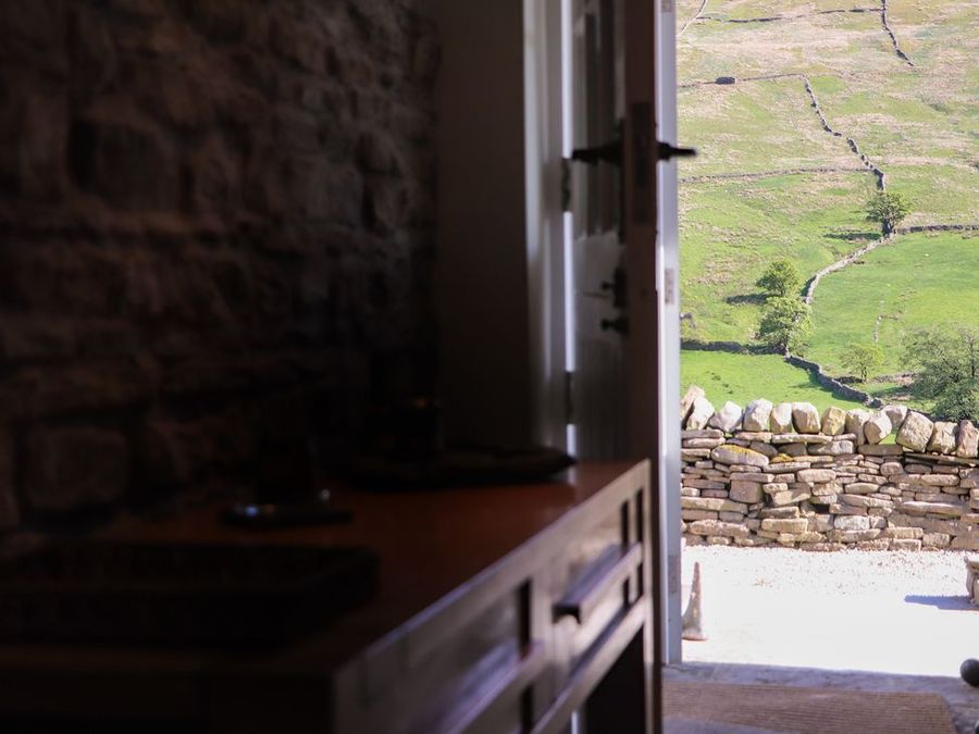 A stone-walled room with wooden furniture looking out through an open door to a stone wall and green fields at Roger Pot in Garsdale near Sedbergh