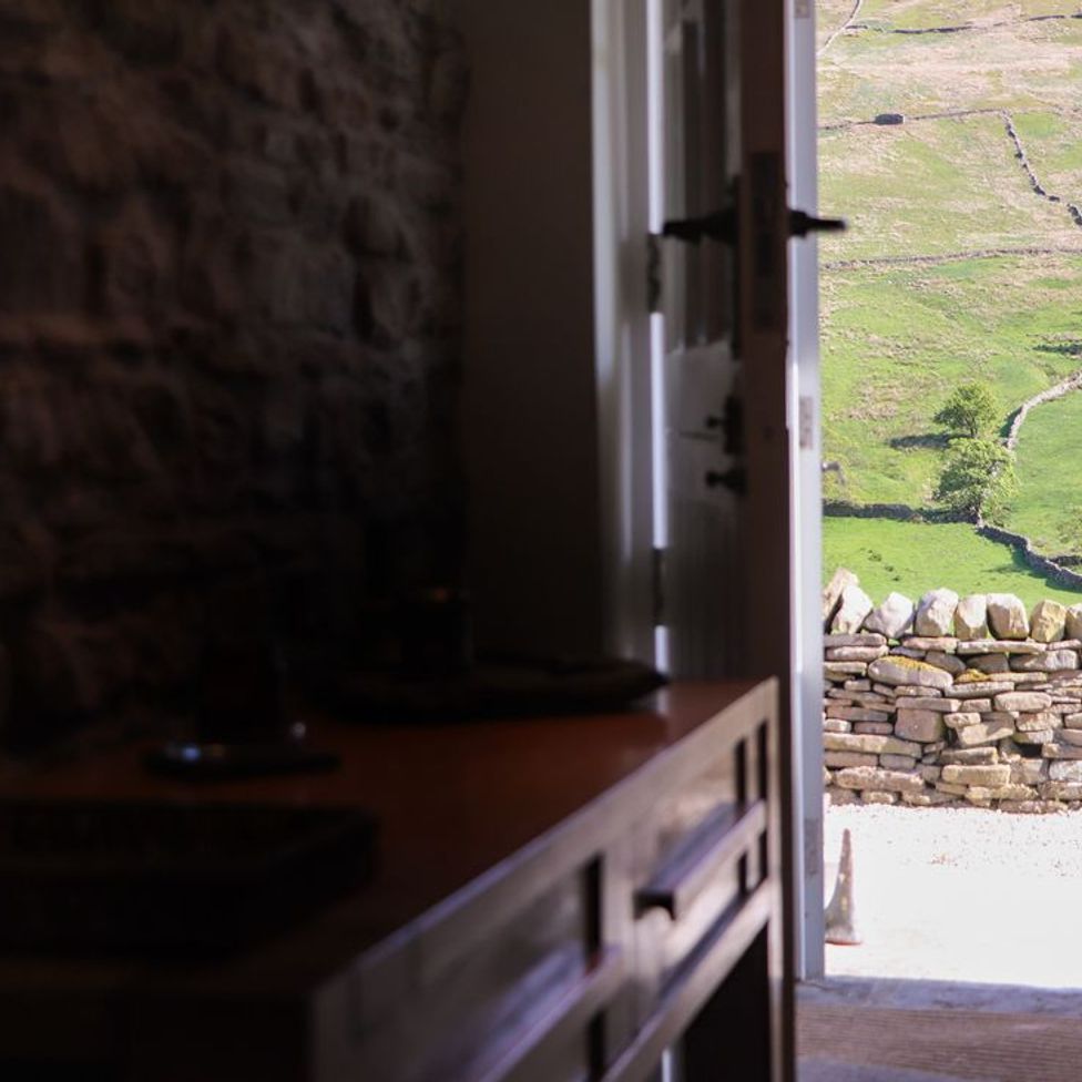 A stone-walled room with wooden furniture looking out through an open door to a stone wall and green fields at Roger Pot in Garsdale near Sedbergh