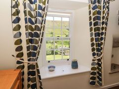 A window with patterned curtains and a bowl and vase on the windowsill overlooking a green hillside at Roger Pot in Garsdale near Sedbergh