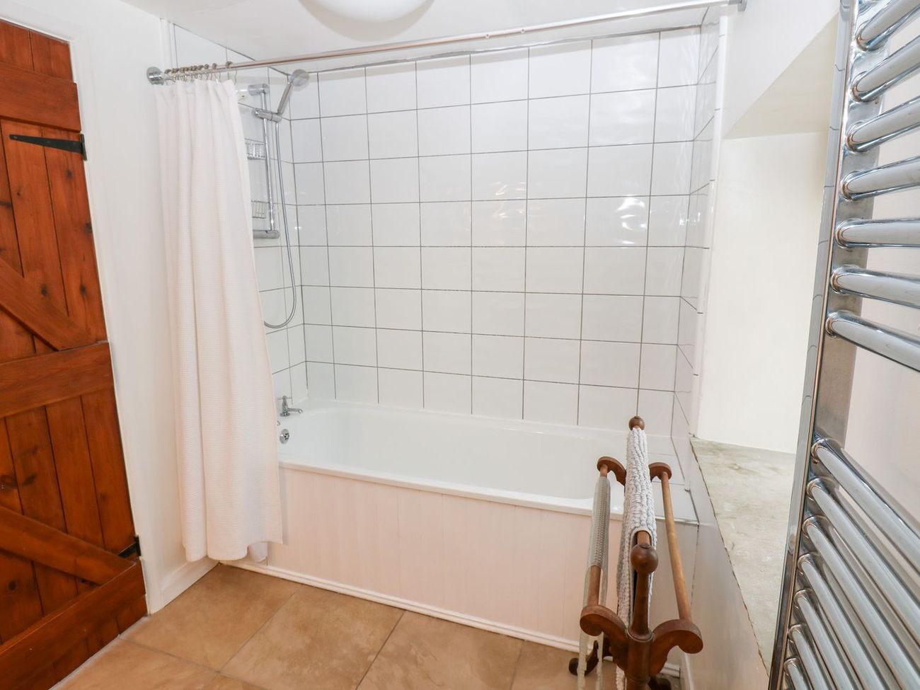The first floor bathroom at Farrier's Cottage, with a bath and shower over it, white tiled walls, a heated towel rail and a stripped pine door