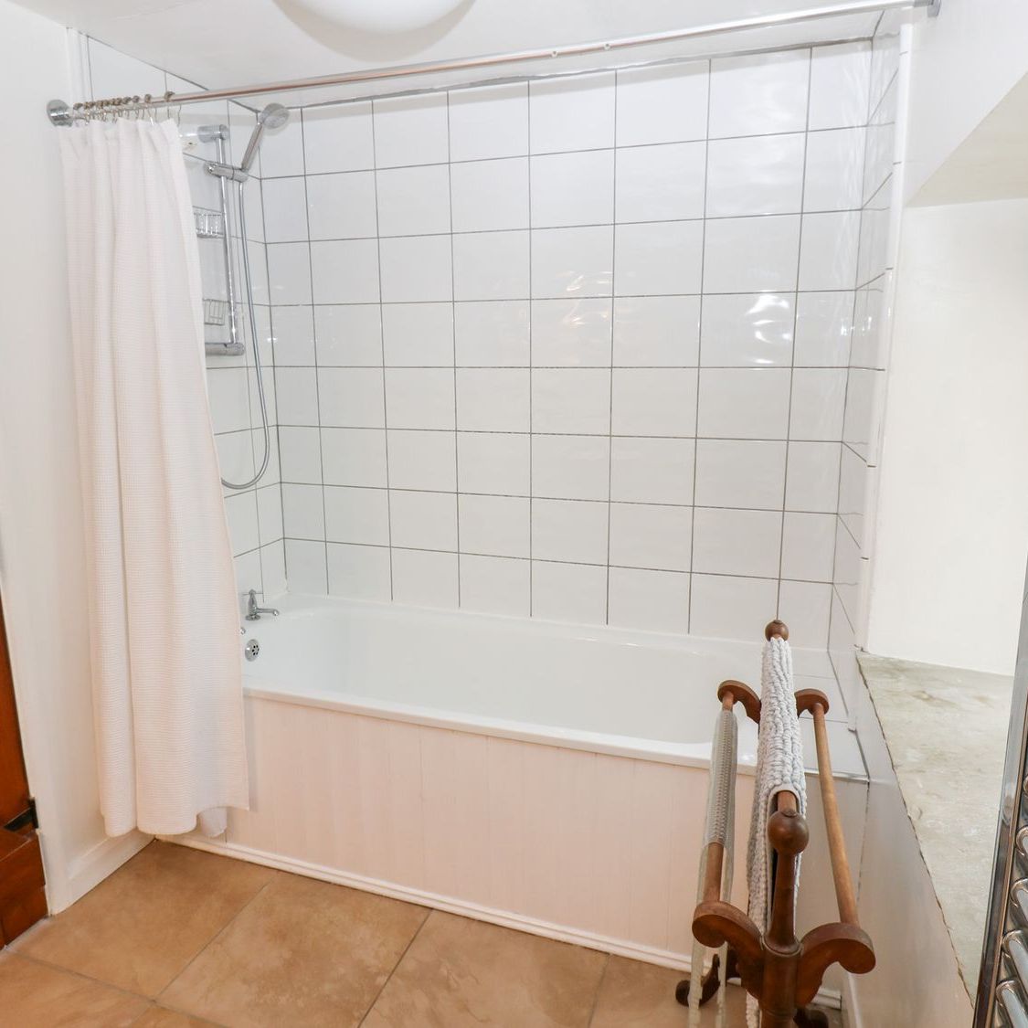 The first floor bathroom at Farrier's Cottage, with a bath and shower over it, white tiled walls, a heated towel rail and a stripped pine door