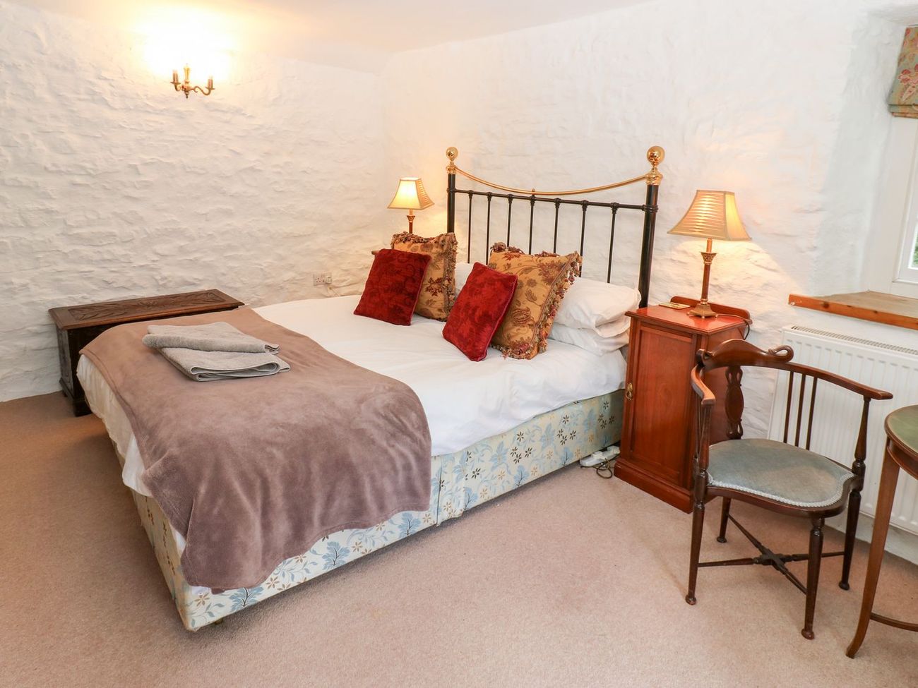 The king-size bedroom at Farrier's Cottage, with a brass and iron bedstead against a whitewashed stone wall, lamps lit on either side
