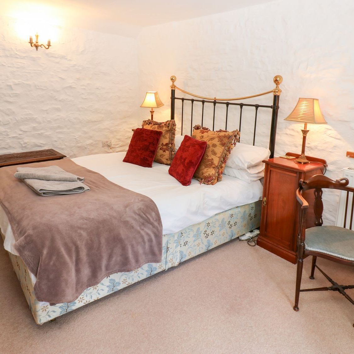 The king-size bedroom at Farrier's Cottage, with a brass and iron bedstead against a whitewashed stone wall, lamps lit on either side