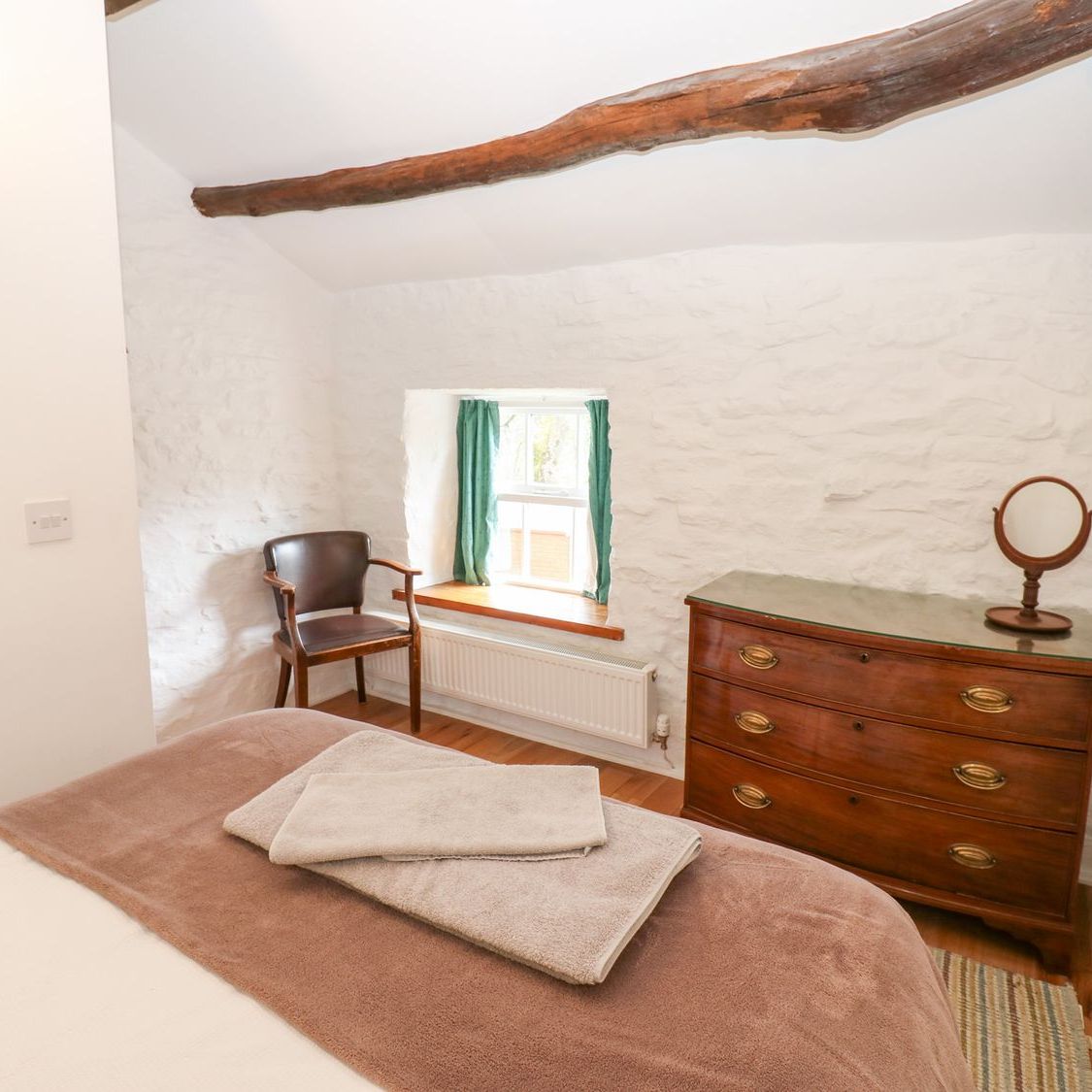 A bedroom at Farrier's Cottage with an exposed roof beam overhead, an antique chest of drawers and towels laid out on the bed