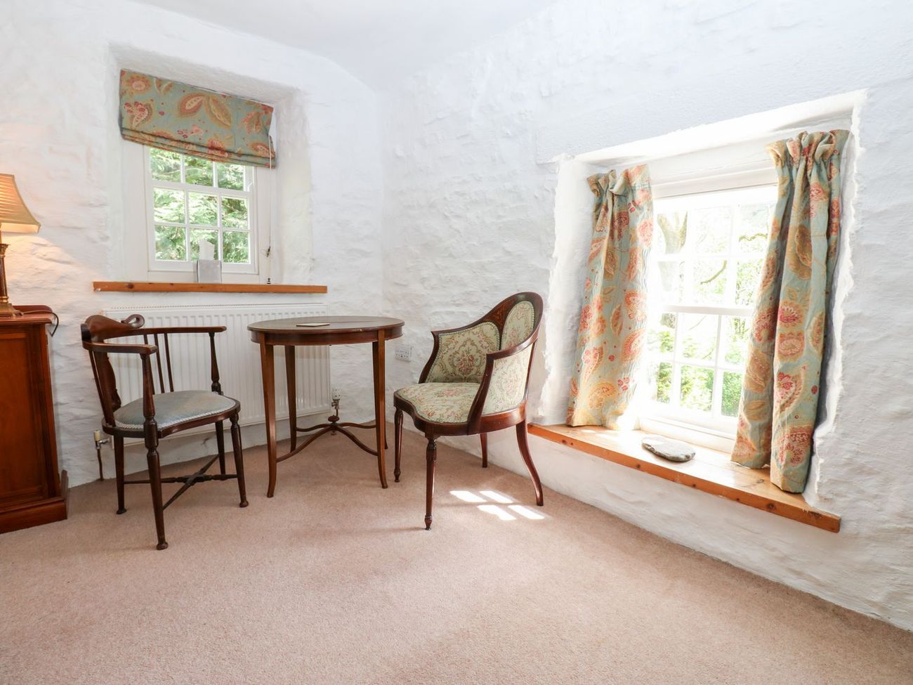 A quiet corner of a bedroom at Farrier's Cottage, with two antique chairs and a round table between two windows looking onto trees