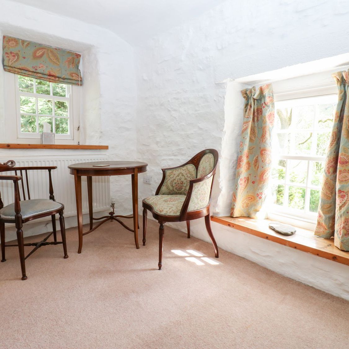 A quiet corner of a bedroom at Farrier's Cottage, with two antique chairs and a round table between two windows looking onto trees