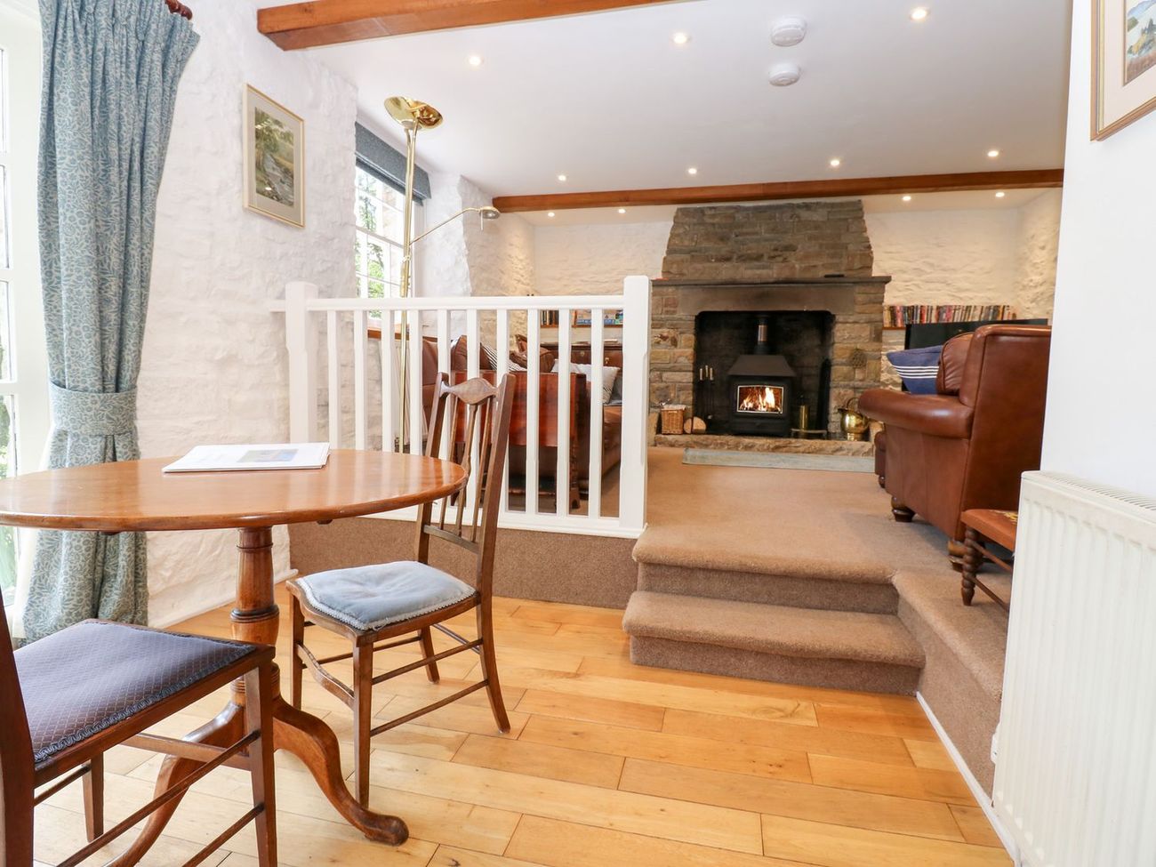 The dining table at Farrier's Cottage, with steps leading up to the sitting room and a fire burning in the stone fireplace beyond