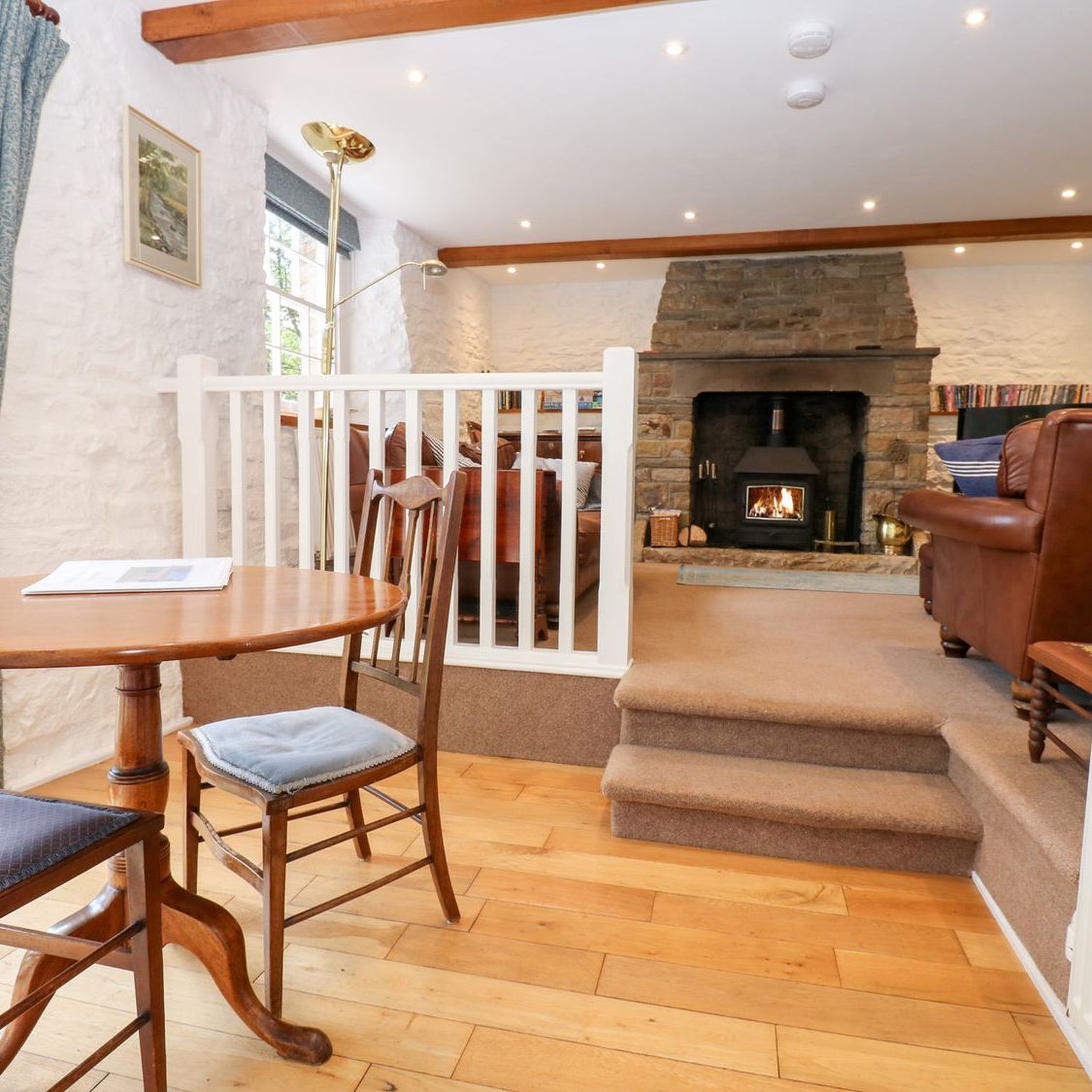 The dining table at Farrier's Cottage, with steps leading up to the sitting room and a fire burning in the stone fireplace beyond
