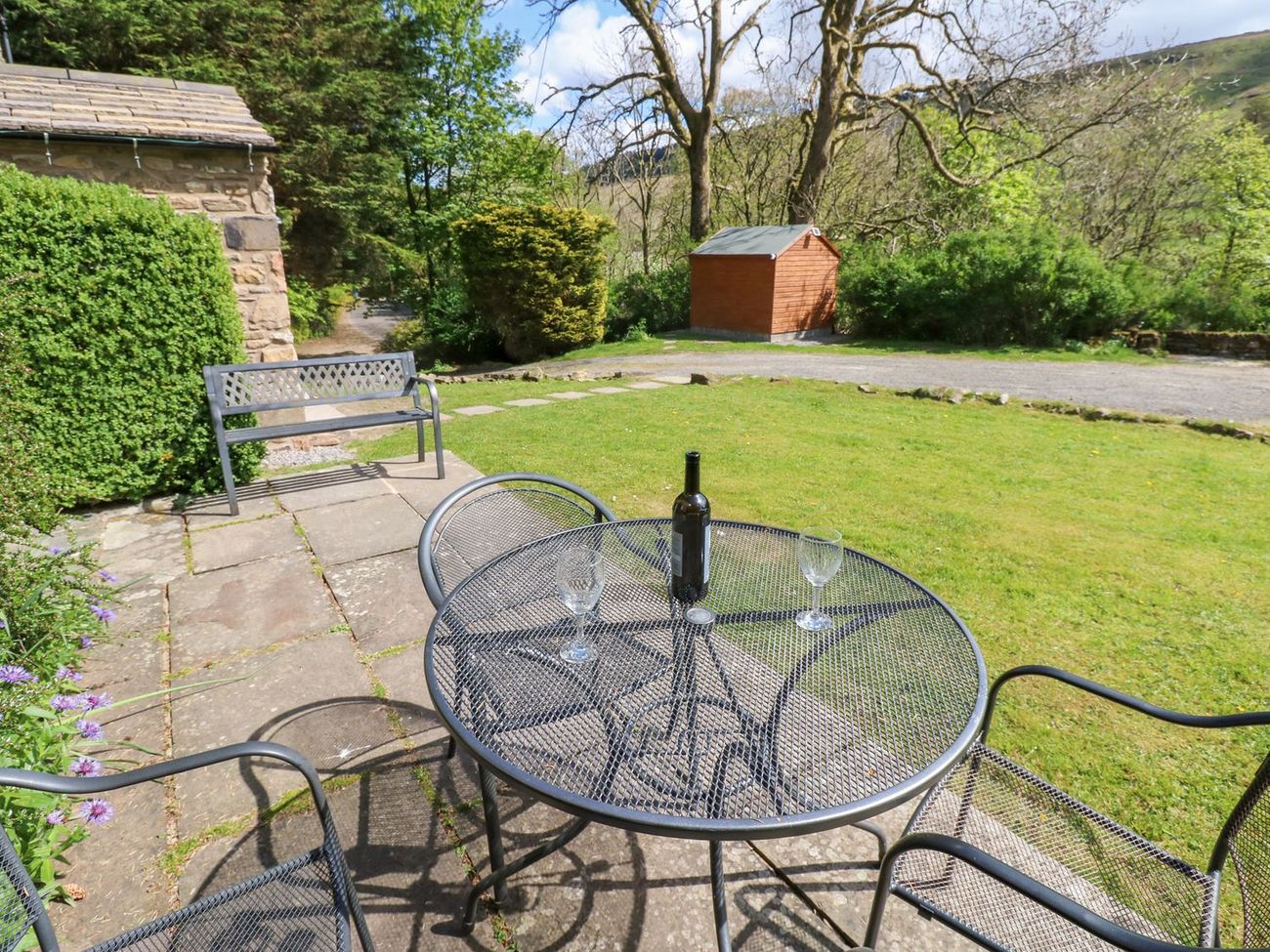 The flagged patio at Farrier's Cottage looking out over the lawn to the fellside, with a garden bench and a table set with a bottle and glasses