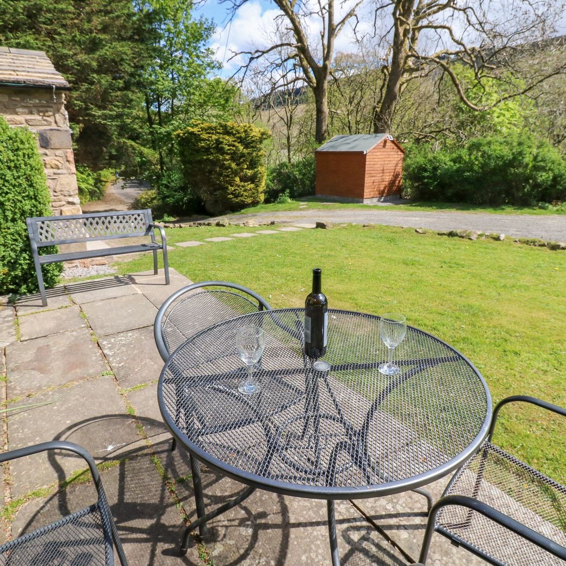 The flagged patio at Farrier's Cottage looking out over the lawn to the fellside, with a garden bench and a table set with a bottle and glasses