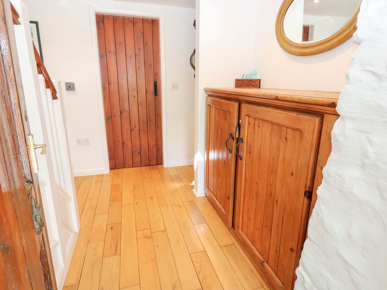 The hallway at Farrier's Cottage, with a wooden floor, a pine cupboard and a gilt mirror on a whitewashed stone wall