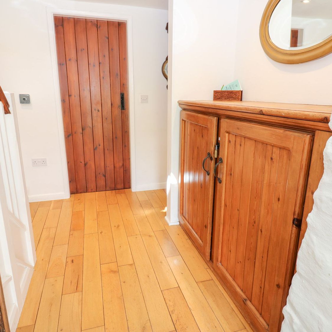 The hallway at Farrier's Cottage, with a wooden floor, a pine cupboard and a gilt mirror on a whitewashed stone wall