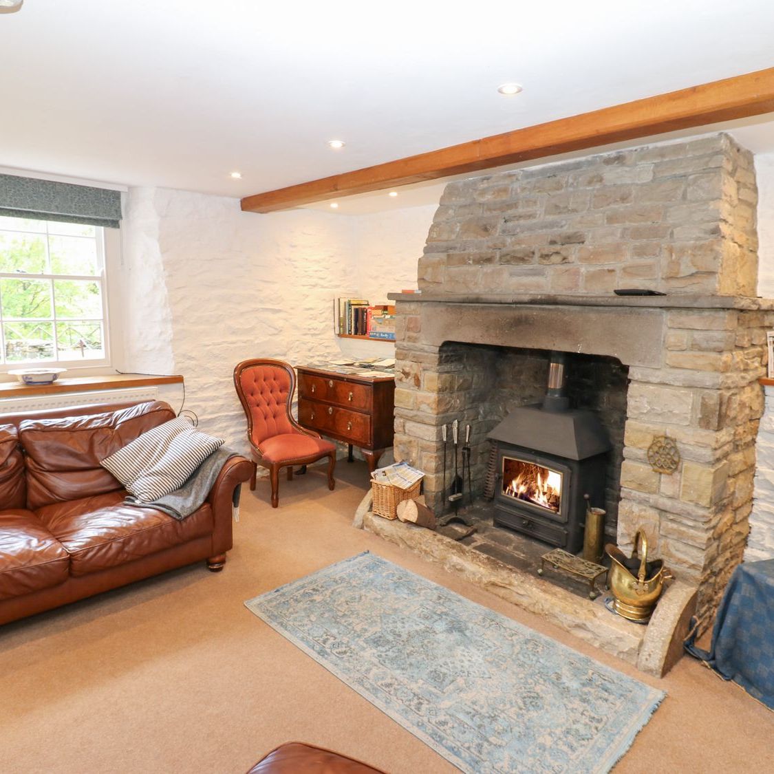 The sitting room at Farrier's Cottage, with a woodburning stove lit in the stone fireplace and a leather sofa beside the window