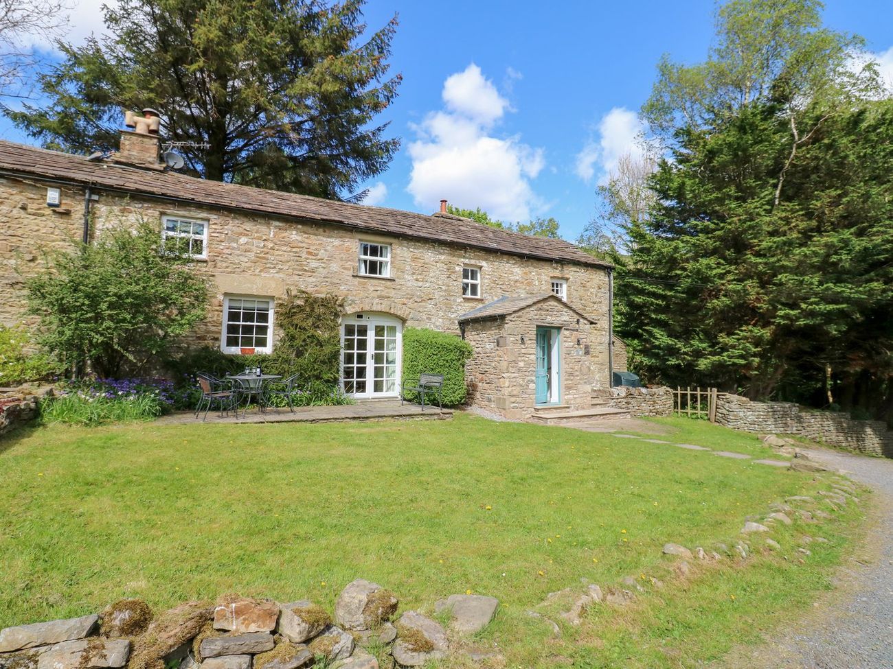 Farrier's Cottage from the garden, a stone cottage with a blue front door, a lawn and a table and chairs on the patio