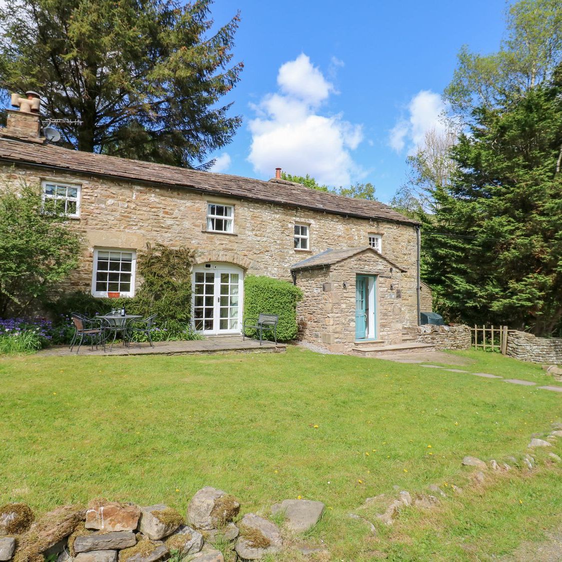 Farrier's Cottage from the garden, a stone cottage with a blue front door, a lawn and a table and chairs on the patio