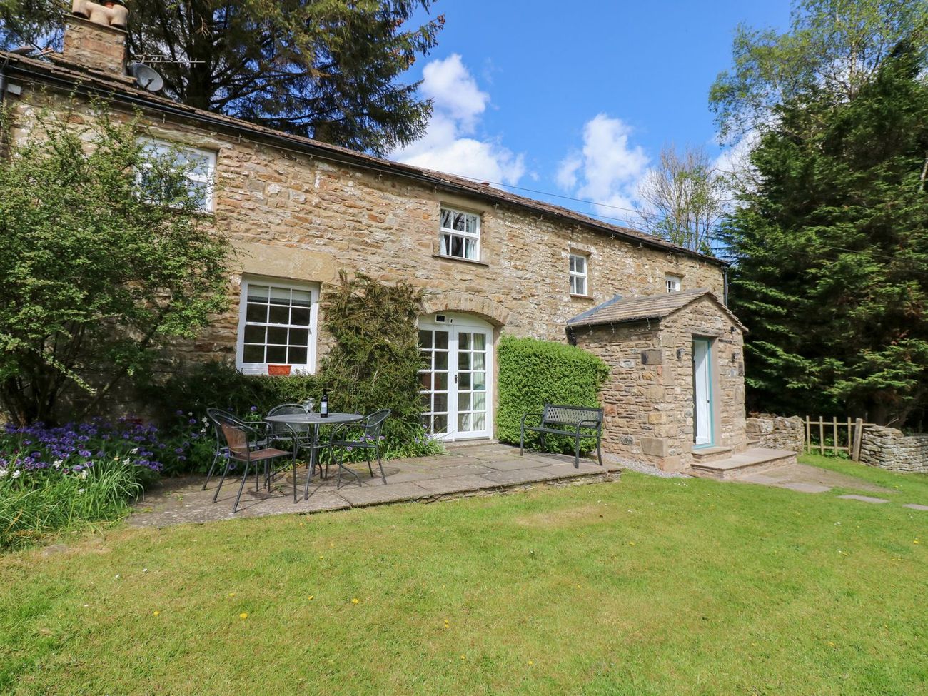 The front of Farrier's Cottage in spring, with French doors onto the patio, a garden bench and flowers in the border