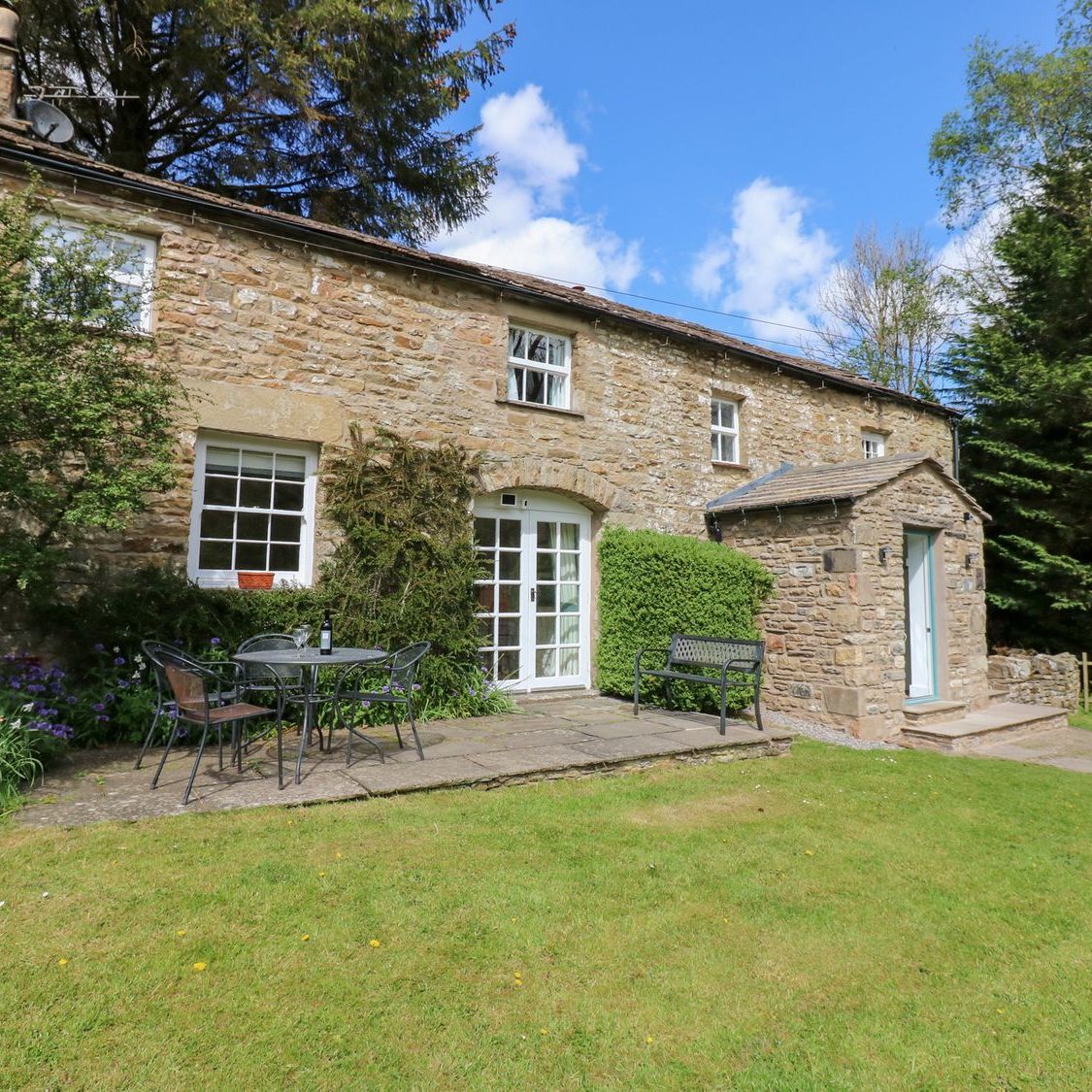 The front of Farrier's Cottage in spring, with French doors onto the patio, a garden bench and flowers in the border