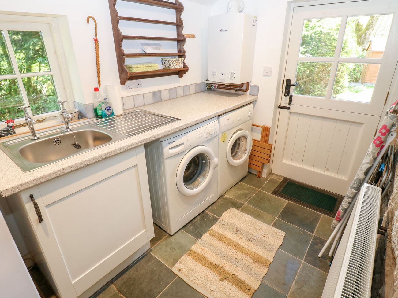 The utility room at Farrier's Cottage, with a washing machine, tumble dryer, sink and a slate floor, and a door out to the garden