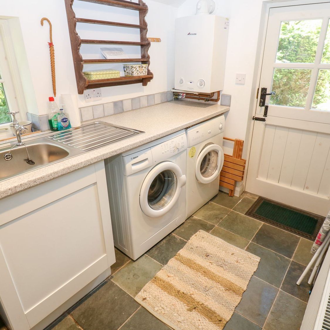 The utility room at Farrier's Cottage, with a washing machine, tumble dryer, sink and a slate floor, and a door out to the garden