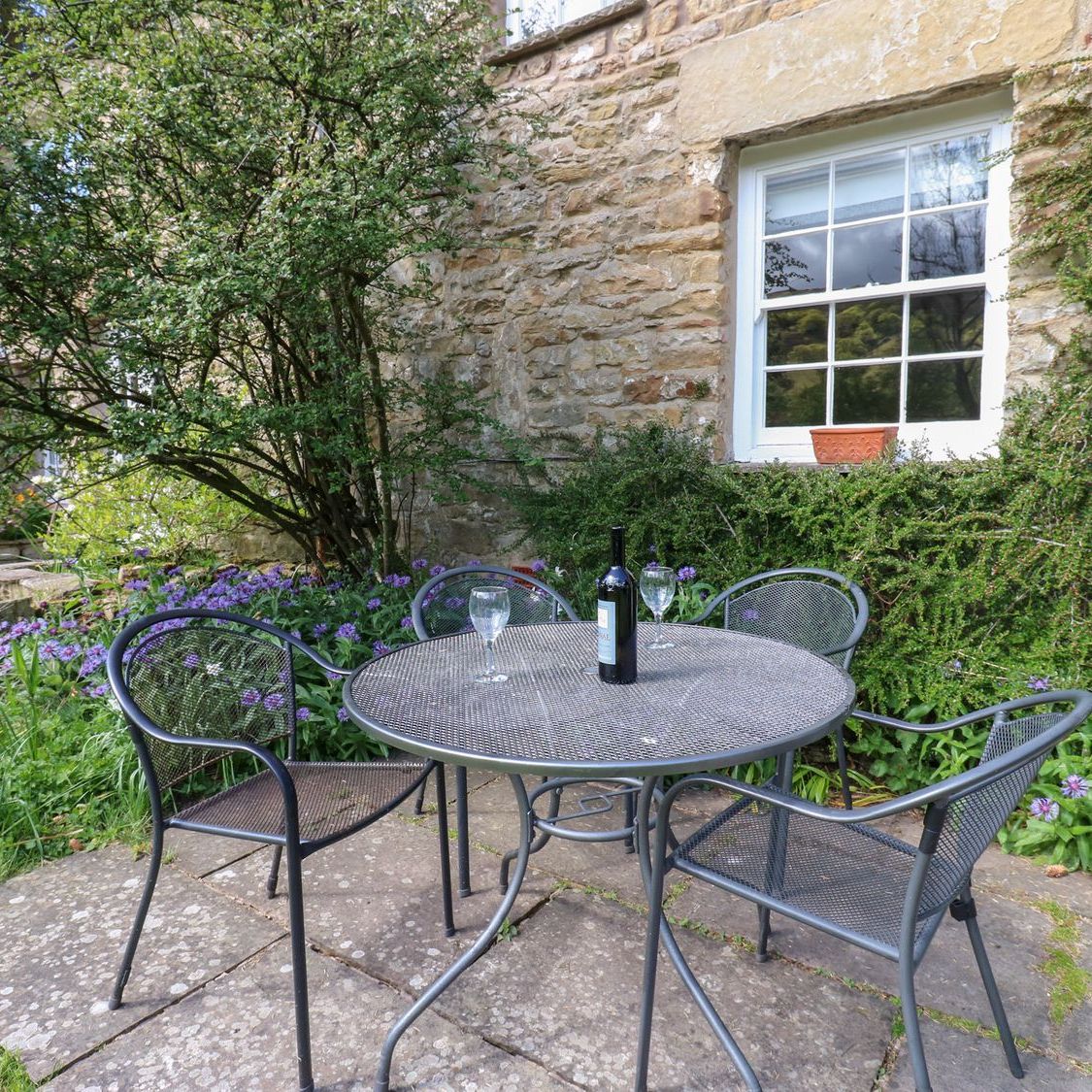 A table and four chairs on the patio at Farrier's Cottage, sheltered by the cottage wall with flowers in the border