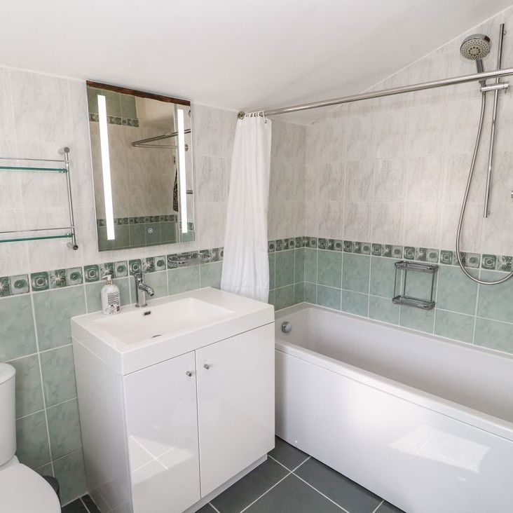 A bathroom at Roger Pot with pale green tiles, a vanity basin, mirror and a bath with shower over