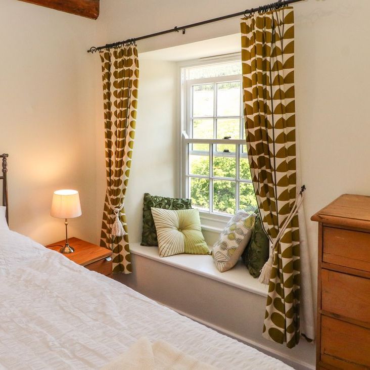 A bedroom at Roger Pot with a metal-framed bed, bedside lamp, cushioned window seat and leaf-patterned curtains