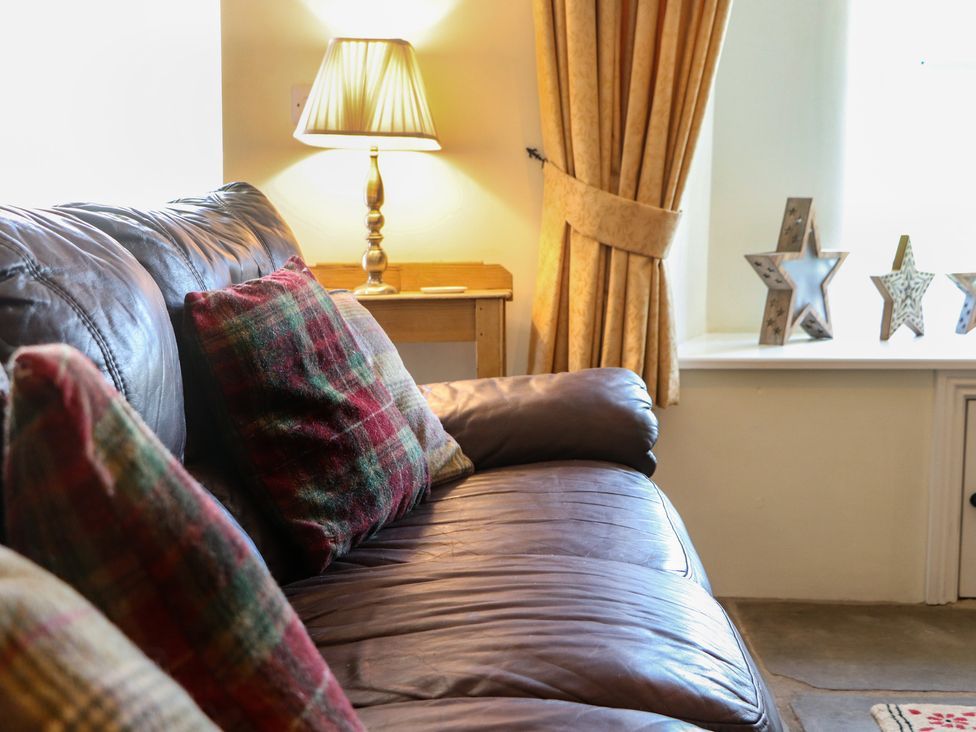 A leather sofa with plaid cushions next to a wooden side table with a lamp and yellow curtains with star decorations on the windowsill at Roger Pot in Garsdale near Sedbergh