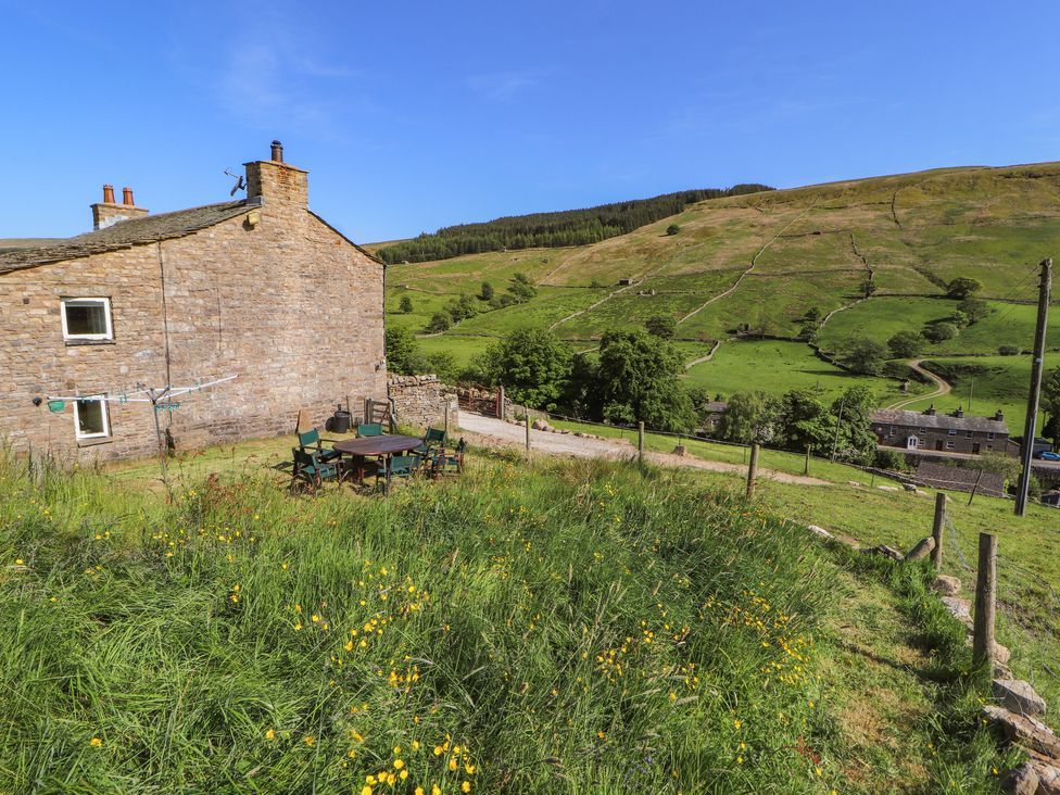 A stone house with a round table and chairs on grass overlooking green hills and fields at Roger Pot in Garsdale near Sedbergh