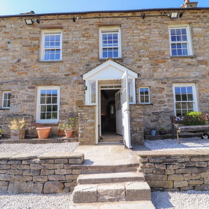 The front of Roger Pot stone house with white sash windows, a porch, potted plants and a cat under the bench