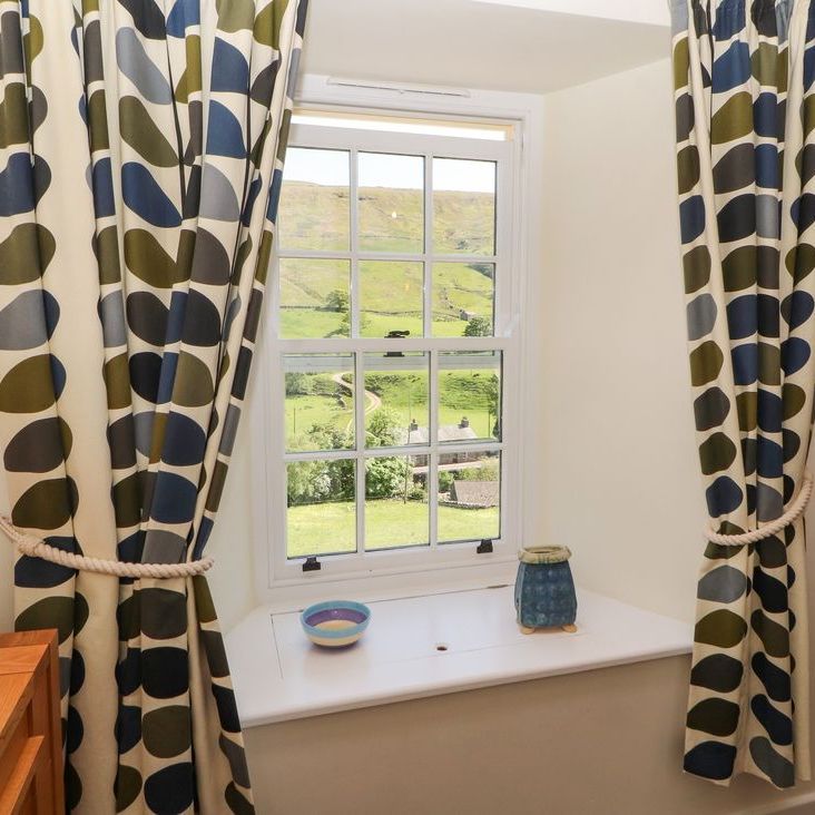 A cottage window at Roger Pot with leaf-patterned curtains, looking out over green fields and a track
