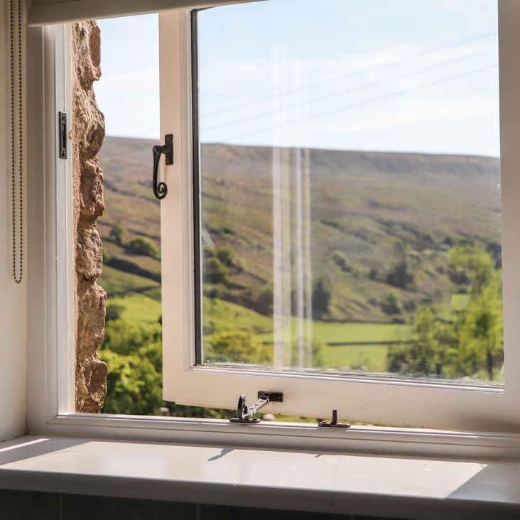 An open cottage window at Roger Pot looking out over green trees, fields and a hillside