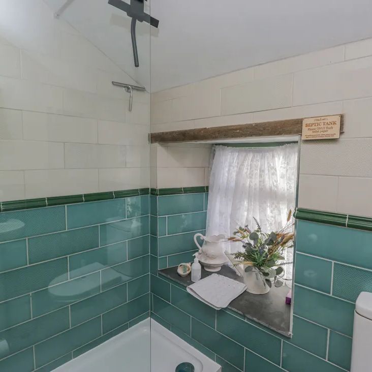 A cottage shower room at The Old Cart House with teal and white tiles, a glass shower and a net-curtained window