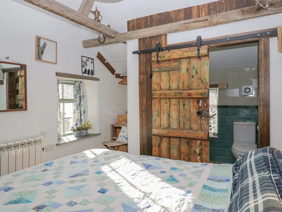 A bedroom with a patterned quilt on the bed a wooden sliding door leading to a bathroom and a window with floral curtains at The Old Cart House in Garsdale near Sedbergh