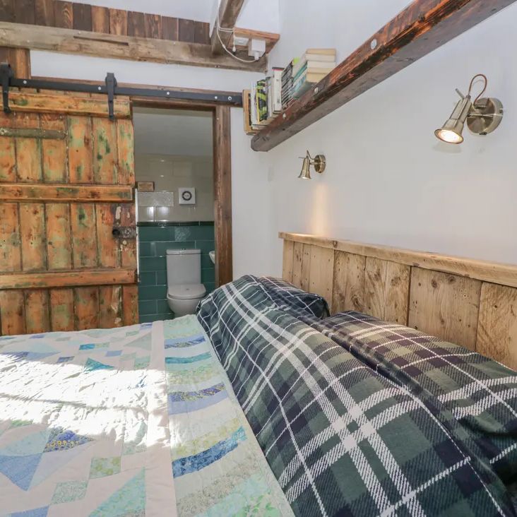 A beamed bedroom at The Old Cart House with a plank-headboard bed, patchwork quilt and sliding barn door to the en-suite